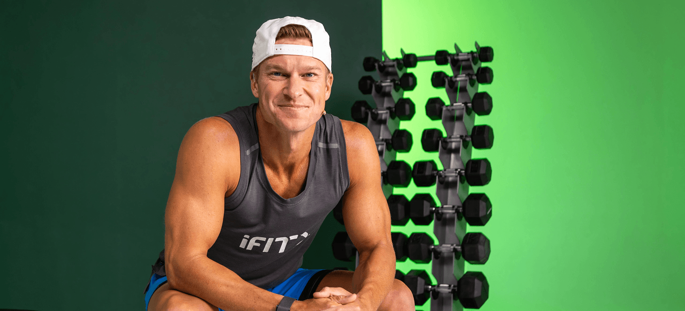 iFIT Trainer John Peel sitting in front of a dumbbell rack, smiling and welcoming the user to a foundational strength training routine.